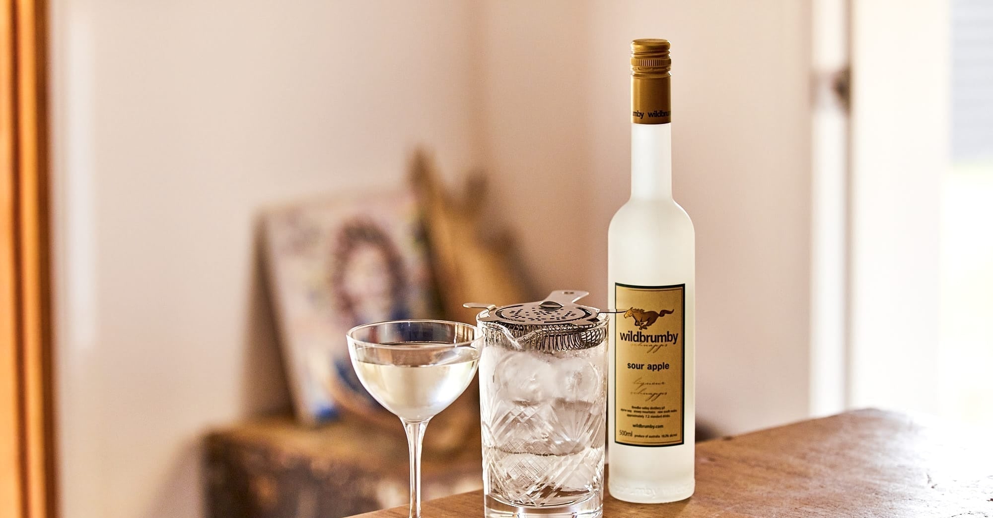 A bottle of Whiskey Aged 12 Years by The Whiskey Company, displayed on a wooden table next to a coupe glass and a rocks glass with ice, in a warm, softly lit indoor setting.