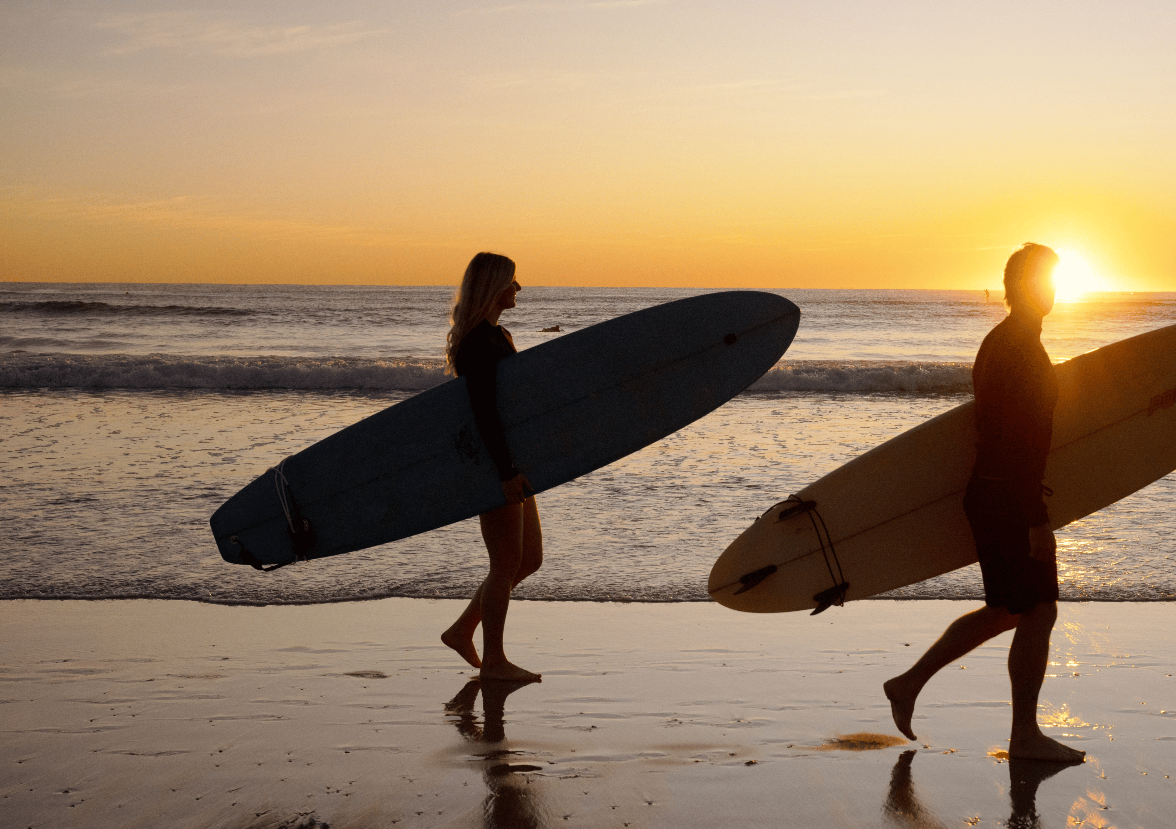 Two surfers carrying surfboards walk along a sandy beach at sunset, silhouetted against the ocean and glowing sun.