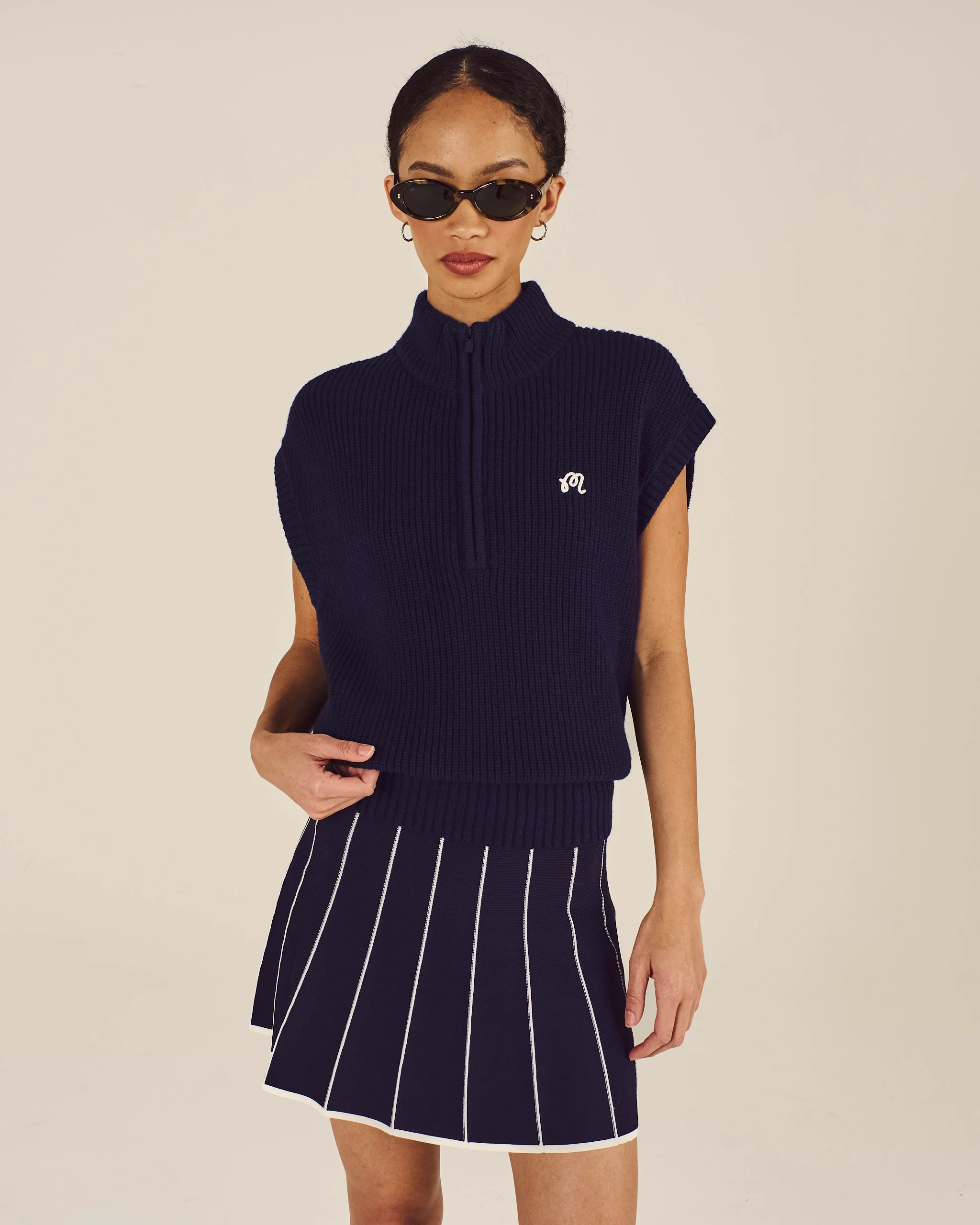 Woman wearing a navy sleeveless sweater and pleated skirt on a beige background